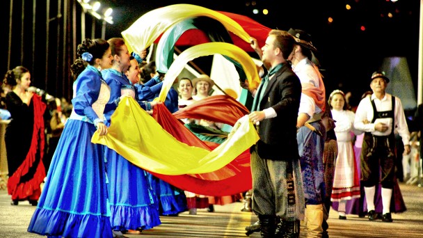 Grupo de casais vestidos com roupas tradicionalistas do Rio Grande do Sul segurando e sacudindo tecidos nas cores amarela, verde e vermelha.